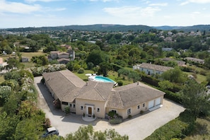 A large, single-story house with a swimming pool, surrounded by lush greenery and other residential buildings.