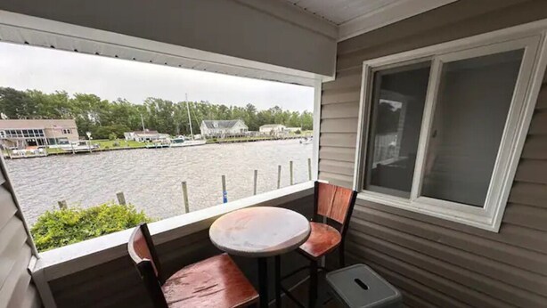 A balcony with a table and two chairs, overlooking a body of water with boats and buildings in the background. - Knot Three- Balcony View of ICW- Dog Friendly! (Coinjock)