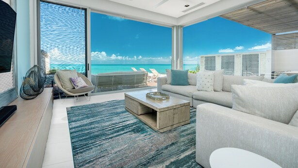 A modern living room with a large window overlooking a beach, a ceiling fan, and a minimalist design. - Long Bay Beach Club (Providenciales)