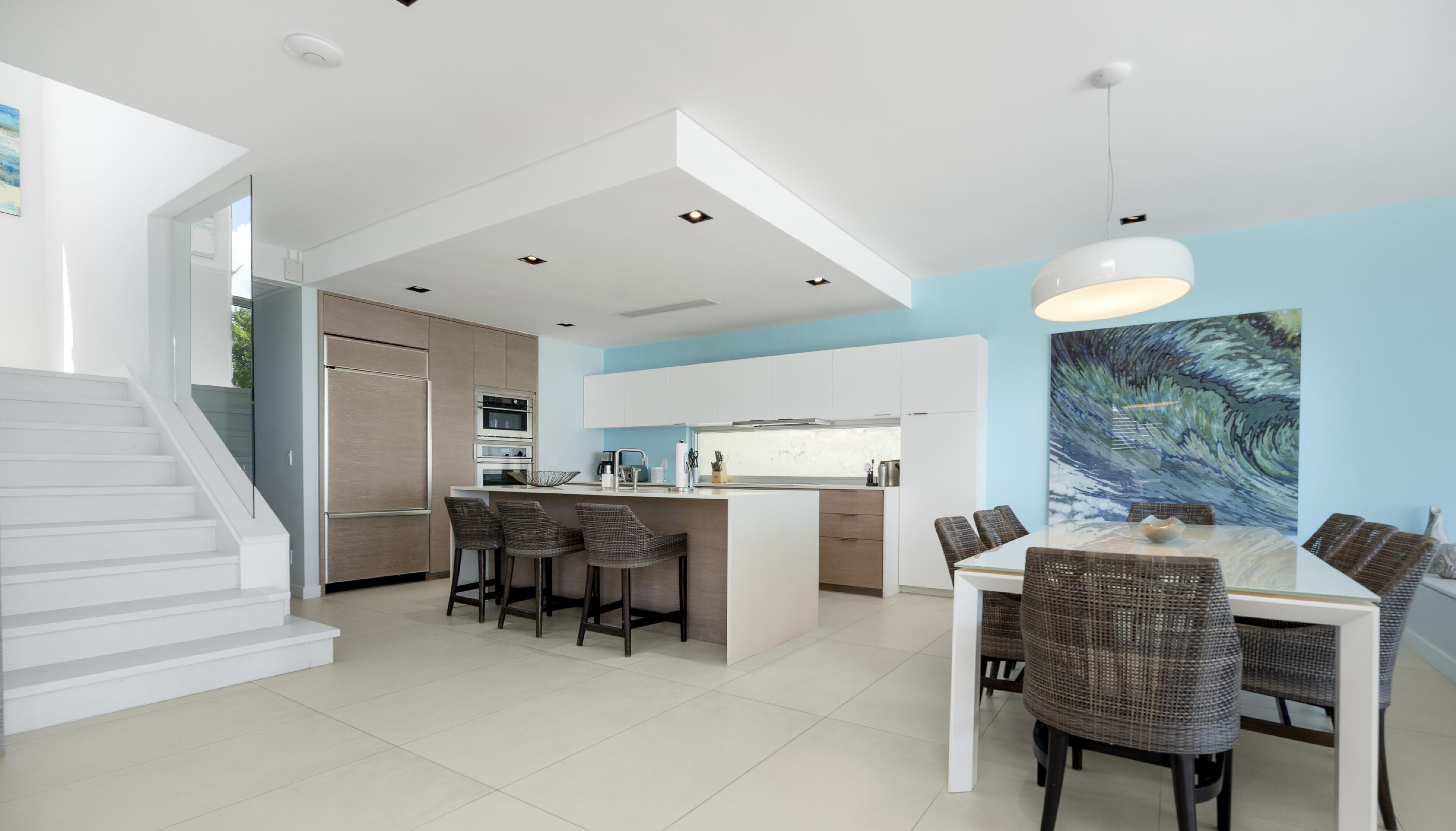 Une cuisine moderne avec des meubles blancs, un îlot central doté de tabourets de bar et un coin repas avec une table blanche et des chaises en osier.
