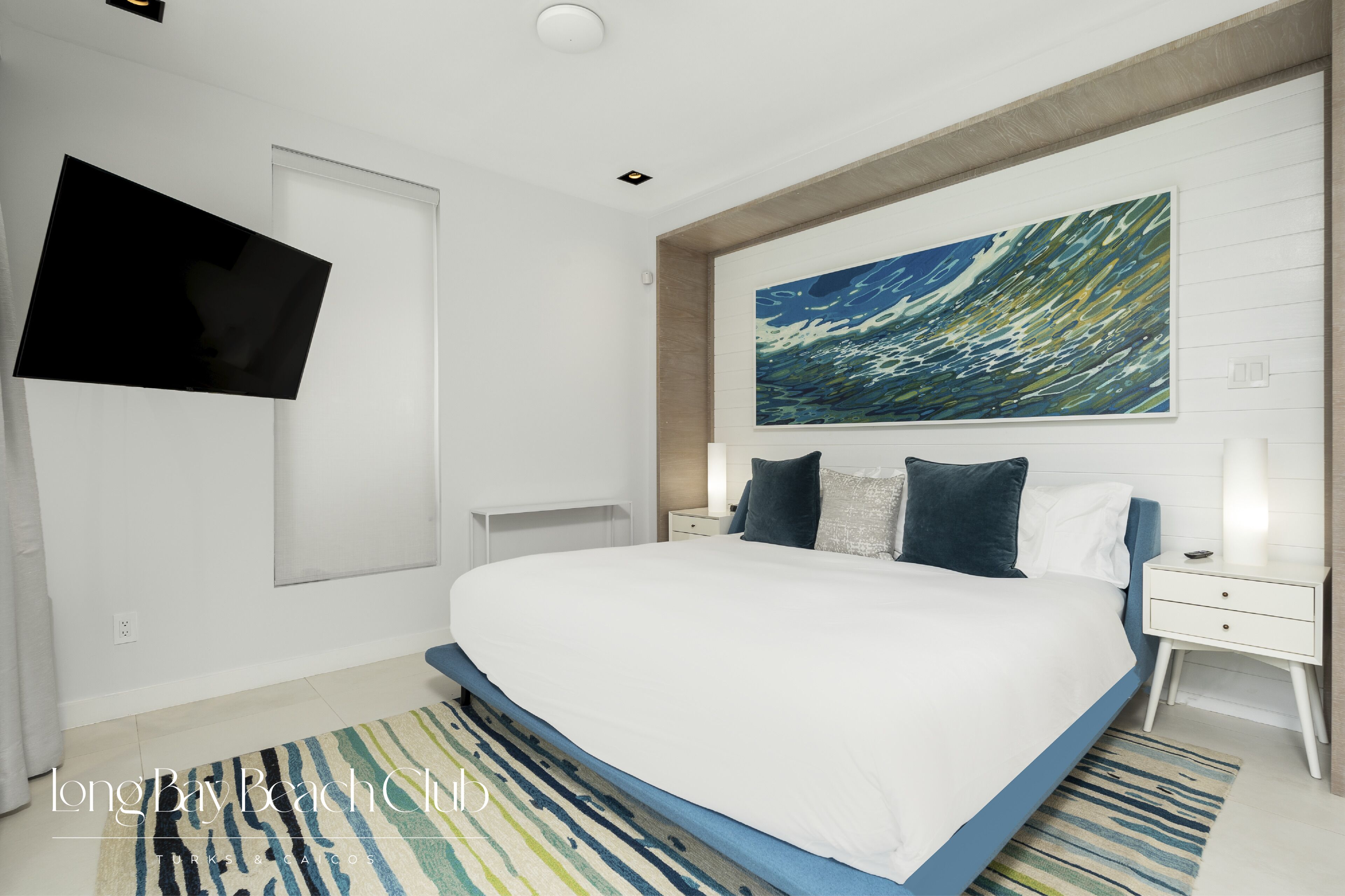 A modern bedroom with a large bed, a mounted TV, a painting, and a nightstand with a lamp.