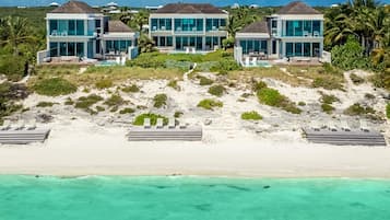 Three villas offer expansive ocean views from large windows, situated on a sandy beach with access via steps.