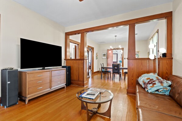 A spacious living room with hardwood flooring, a large flat-screen TV, a wooden cabinet, a glass coffee table, and a brown leather sofa. - The O Place - Center of town and close to everything! (Galveston)