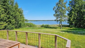 Une terrasse en bois avec une rambarde en métal, donnant sur un lac, et entourée d’arbres et d’herbe.