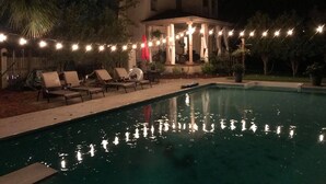 A swimming pool with lounge chairs and string lights, surrounded by trees and a building. - French Tudor in Quiet Aiken County (Aiken)