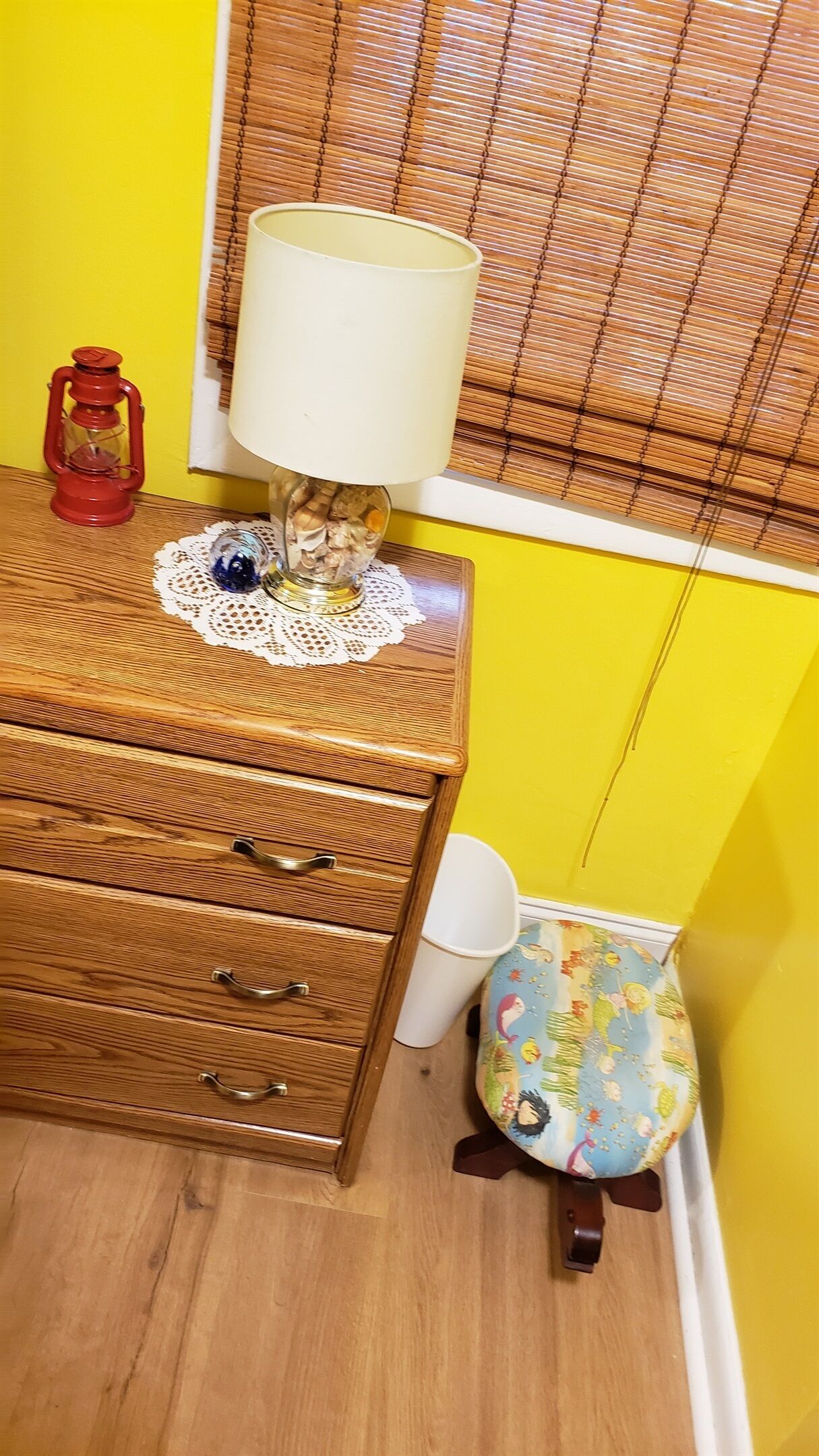 A wooden dresser with drawers, a white lamp on a doily, a red lantern, and a colorful bag against a yellow wall.
