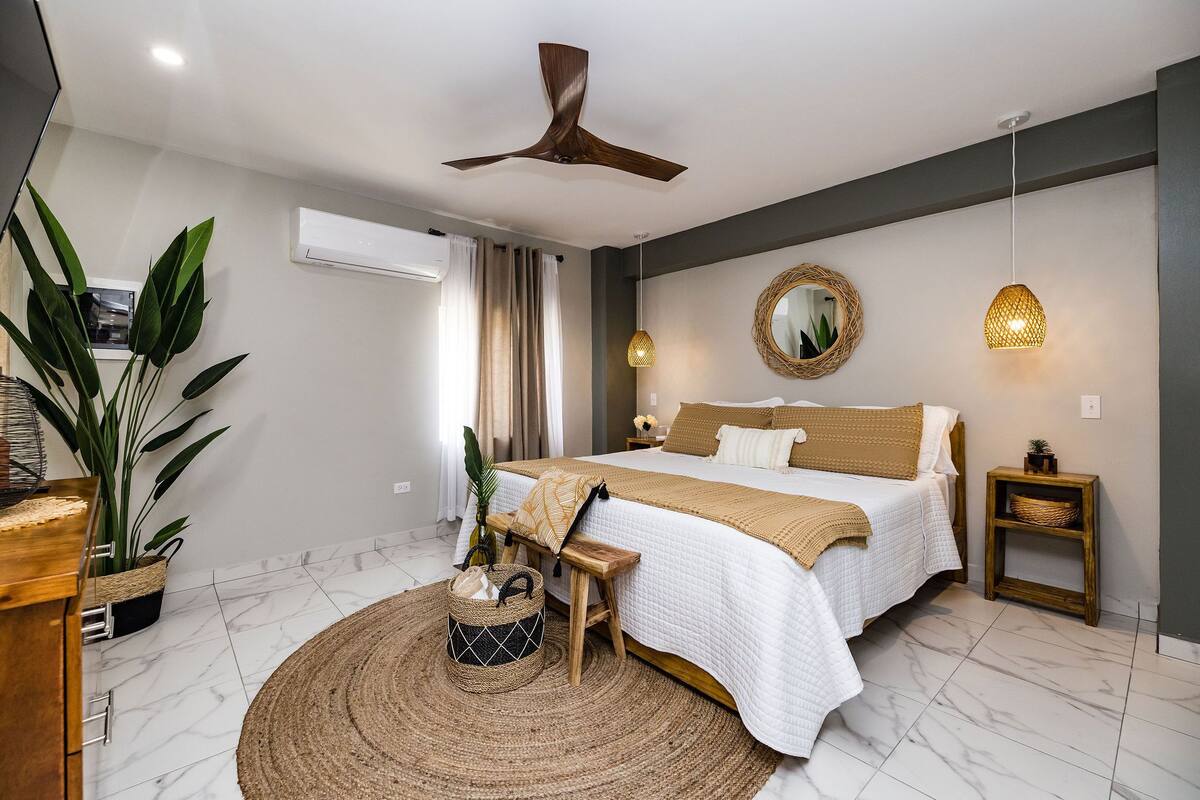 A modern bedroom with a bed, bedside tables, a ceiling fan, a large mirror, and a potted plant.