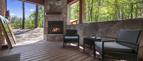 A wooden deck with a stone fireplace, wicker chairs, and a view of a forest.