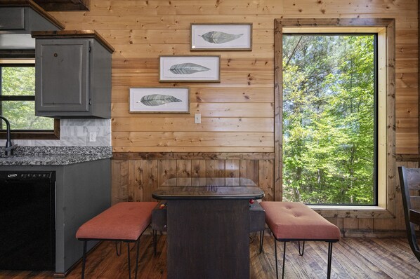 Interior de una cabaña de madera con zona de cocina, una mesa con bancos y arte con hojas enmarcadas en la pared.