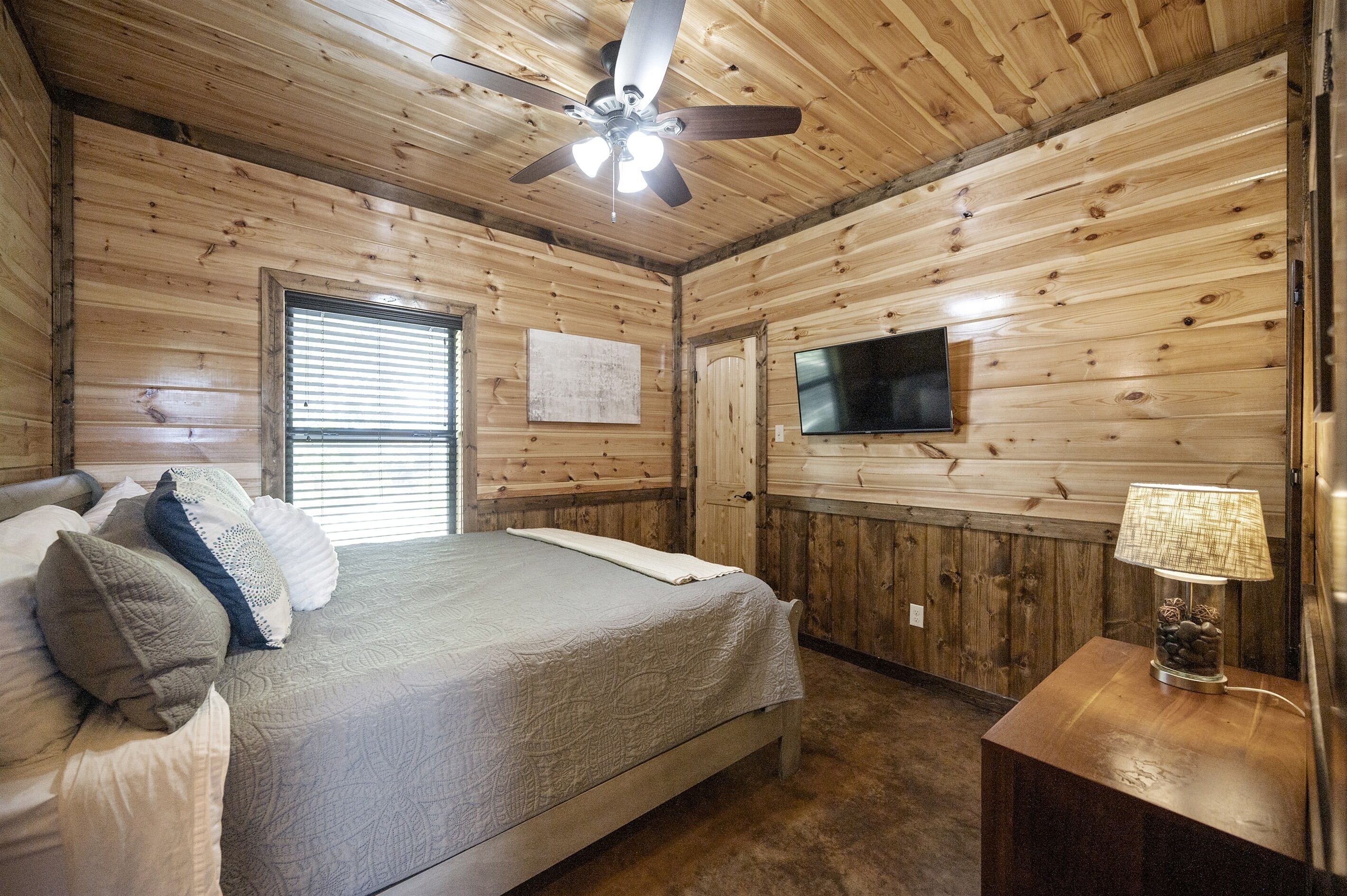 A wooden-paneled bedroom with a bed, a ceiling fan, a TV, and a nightstand with a lamp.