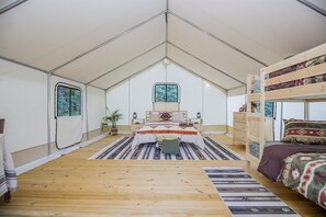 Ein Zeltraum mit einem zentralen Bett, einem Schlafplatz in Form eines Etagenbettes rechts und einem kleinen Tisch mit Stuhl. An der linken Wand befinden sich zwei Fenster und eine Tür.