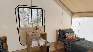 A tent with two beds, a shelf with towels, a small table, and a window with a view of trees.