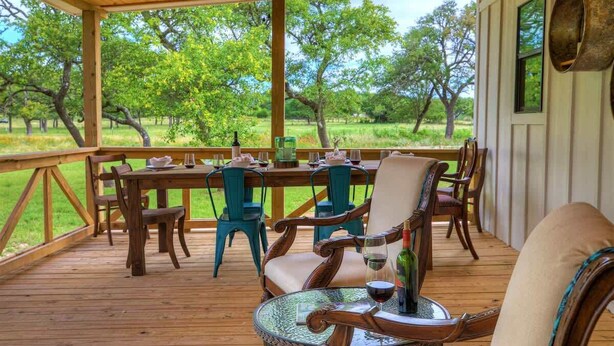 A covered outdoor dining area with a wooden table and chairs, a glass table, and a view of trees and grass. - 3 Acres of Luxury and Views in Fred Wine Country! (Fredericksburg)