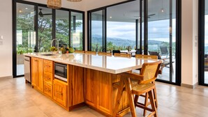 A modern kitchen with a large island, wooden cabinets, and large windows offering a view of the outdoors. - Modern Luxury with Expansive Ocean View and Covered Patio -Private Waterfall! (Uvita)