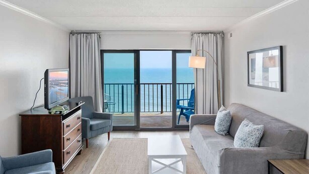A room with a sofa, armchair, TV, and a view of the sea through large windows. - Oceanfront Condo with Dual Views (Ocean City)