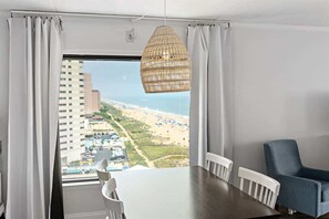 Een kamer met een groot raam, een eettafel, stoelen, een hanglamp en een blauwe stoel.