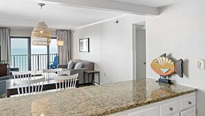 A modern kitchen with granite countertops, a dining area with white chairs, a sofa, and a balcony with a view. - Oceanfront Condo with Dual Views (Ocean City)