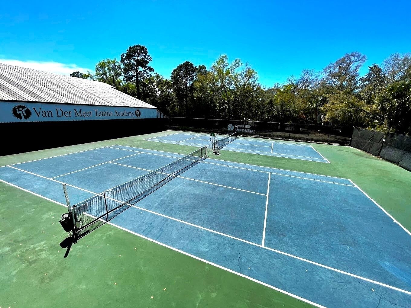 Cancha de tenis con red en el centro, rodeada de árboles y un edificio con el letrero 'Van Der Meer Tennis Academy'.