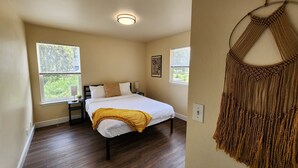 Un dormitorio con una cama, dos mesitas de noche, una mesita auxiliar, un macramé en la pared y dos ventanas con persianas.