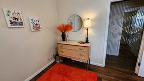 Una habitación con piso de madera, pared color beige, un pequeño aparador beige con cajones, un espejo, una lámpara y dos cuadros colgados en la pared.