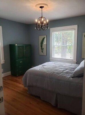 A bedroom with a bed, a chandelier, a window with blinds, and a green cabinet. - Sunny Virginia Equestrian Cottage (Aiken)