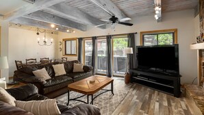 Ein geräumiges Wohnzimmer mit großem Fernseher, Ledersofas, Holzboden und Deckenventilator.