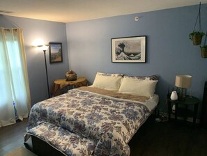 Een slaapkamer met een bed, nachtkastje, lamp en een lijst met een kunstwerk aan de muur.