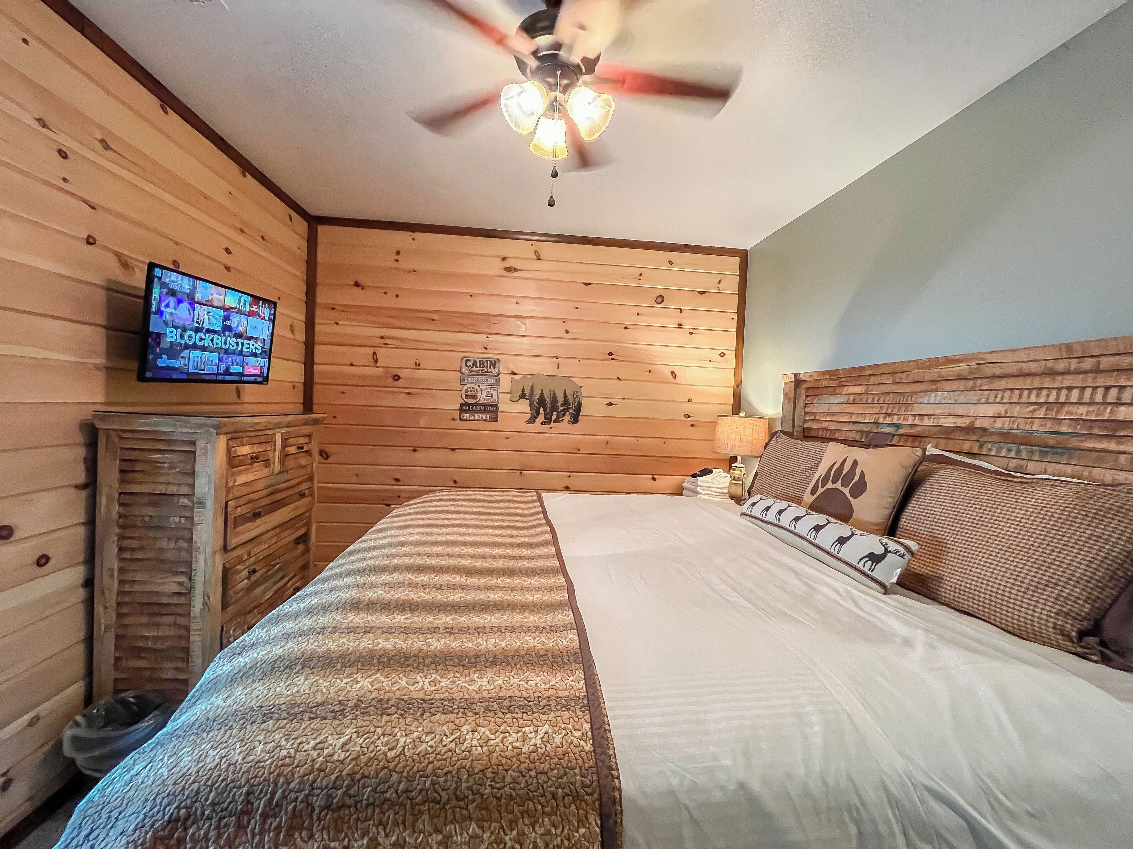 A room with a wooden wall, a bed with patterned bedding, a television on the wall, and a ceiling fan.