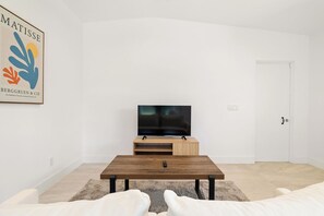 Un salon minimaliste avec un téléviseur à écran plat posé sur un meuble en bois, un canapé blanc et une œuvre d’art encadrée accrochée au mur.