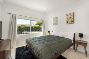 A bedroom with a bed, a TV, a bedside table, a window with curtains, and a view of a swimming pool.