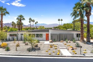 Une maison moderne à deux étages, dotée d’un jardin de style désertique, de palmiers et d’une chaîne de montagnes en arrière-plan.