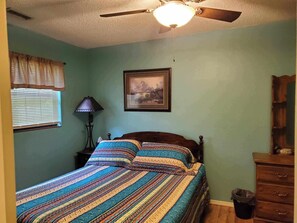 Ein Schlafzimmer mit einem Bett, einem Deckenventilator, einem Fenster mit Jalousien, einer Lampe, einem gerahmten Bild an der Wand und einer Kommode.