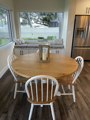 A round wooden dining table with white chairs, a candle centerpiece, and a view of a backyard through large windows.