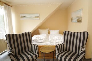 A room with a bed, two striped armchairs, a small round table, and two paintings on the wall.