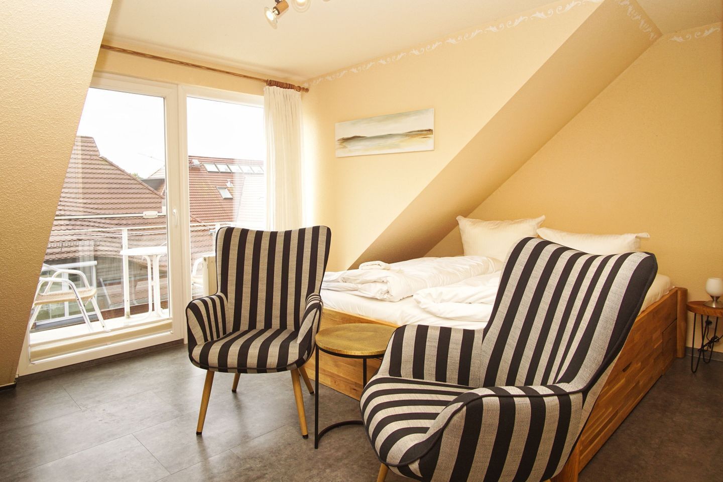 A bedroom with a bed, two striped armchairs, a small table, and a window with a view of buildings.