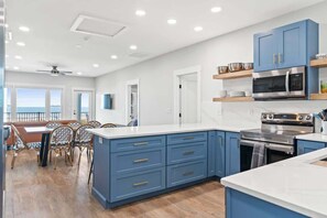 A modern kitchen with blue cabinets, a stainless steel oven, and a dining area with a view. - Cake by the Ocean by Brunswick Vacation Rentals: Fully Renovated Oceanfront Home (Holden Beach)
