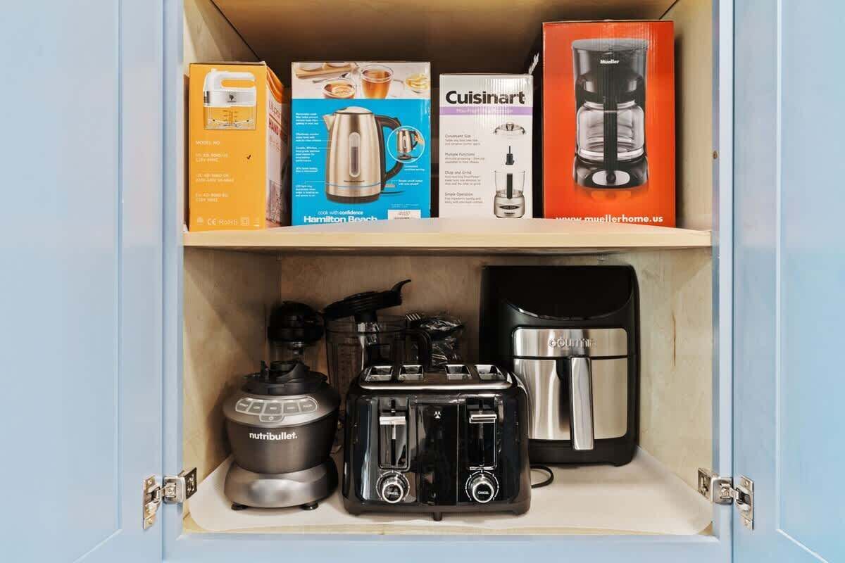 Un meuble de cuisine contenant une machine à café de marque Hamilton Beach, un robot culinaire de marque Cuisinart, un mixeur NutriBullet et un grille-pain.