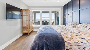 A bedroom with a bed, a ceiling fan, a TV, a bookshelf, and a view of the outdoors. - Cake by the Ocean by Brunswick Vacation Rentals: Fully Renovated Oceanfront Home (Holden Beach)