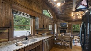A rustic kitchen with wooden cabinets, a granite countertop, a built-in dishwasher, and a stone fireplace. - Creekside Cabin on Private Acreage (Banner Elk)