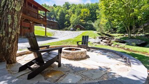 A stone patio with a fire pit, wooden deck chairs, and a view of a lodge with a balcony. - Creekside Cabin on Private Acreage (Banner Elk)