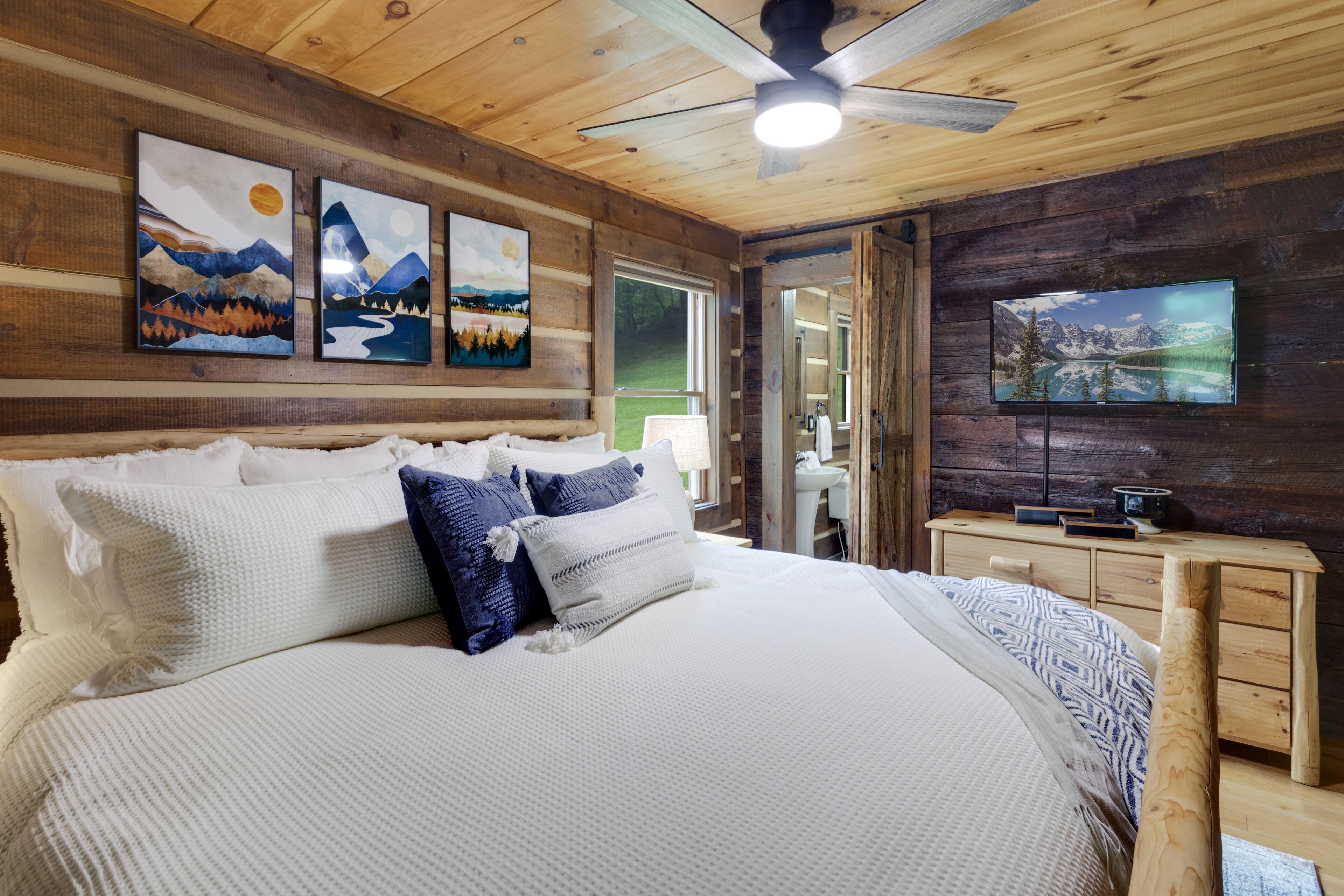 Ein Schlafzimmer einer Holzhütte mit einem Bett, Kissen, einem Deckenventilator, Kunstwerken an den Wänden und einem Fernseher auf einer Kommode.