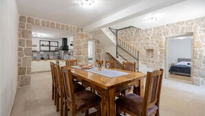 Un vaste espace de salle à manger avec une table et des chaises en bois, une cuisine à l’arrière-plan et un escalier en pierre menant à un niveau supérieur.