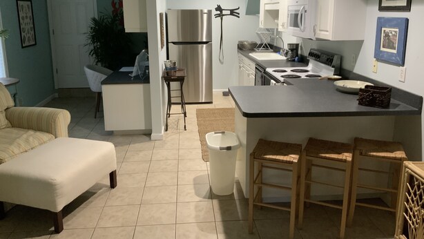 Una cucina moderna con mobili bianchi, un frigorifero in acciaio inossidabile e un piano di lavoro nero. C'è un'area pranzo con un divano bianco e un piccolo tavolo.