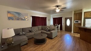 Uma sala de estar com sofá seccional, mesa de centro, luminária, ventilador de teto, área de jantar com mesa e cadeiras, e cozinha com armários de madeira e eletrodomésticos.