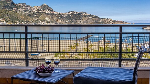 A balcony with a table, two wine glasses, and a bowl of grapes. The view includes a beach, a mountain, and the sea. - Rosal 3-Giardini Naxos holiday home (Giardini Naxos)