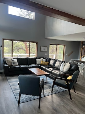 A modern living room with a sectional sofa, a coffee table, and a chair. There are large windows providing natural light and a view of trees.
