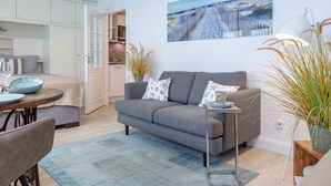 Ein modernes Wohnzimmer mit einem grauen Sofa, einem kleinen Tisch mit einer Lampe und einem Bild von einer Strandlandschaft an der Wand.