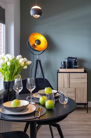 Une table à manger dressée avec des assiettes, des verres et des pommes, un vase de fleurs blanches, une lampe et une machine à café posée sur un meuble.