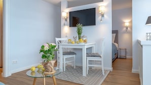 A modern living room with a glass coffee table, white dining table, and chairs. A flat-screen TV is mounted on the wall above the dining area. - 1-zimmerwohnung in Direkter Nähe zum Weitläufigen, Feinsandigen Strand - Andreas-dirks-straße 1 (Sylt)