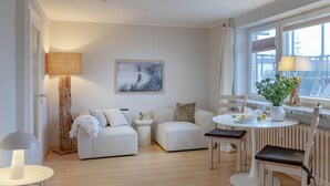 A modern living room with a sofa, a small table, and a dining area with chairs. There is a window with a view of a building outside. - 1-zimmerwohnung in Direkter Nähe zum Weitläufigen, Feinsandigen Strand - Andreas-dirks-straße 1 (Sylt)
